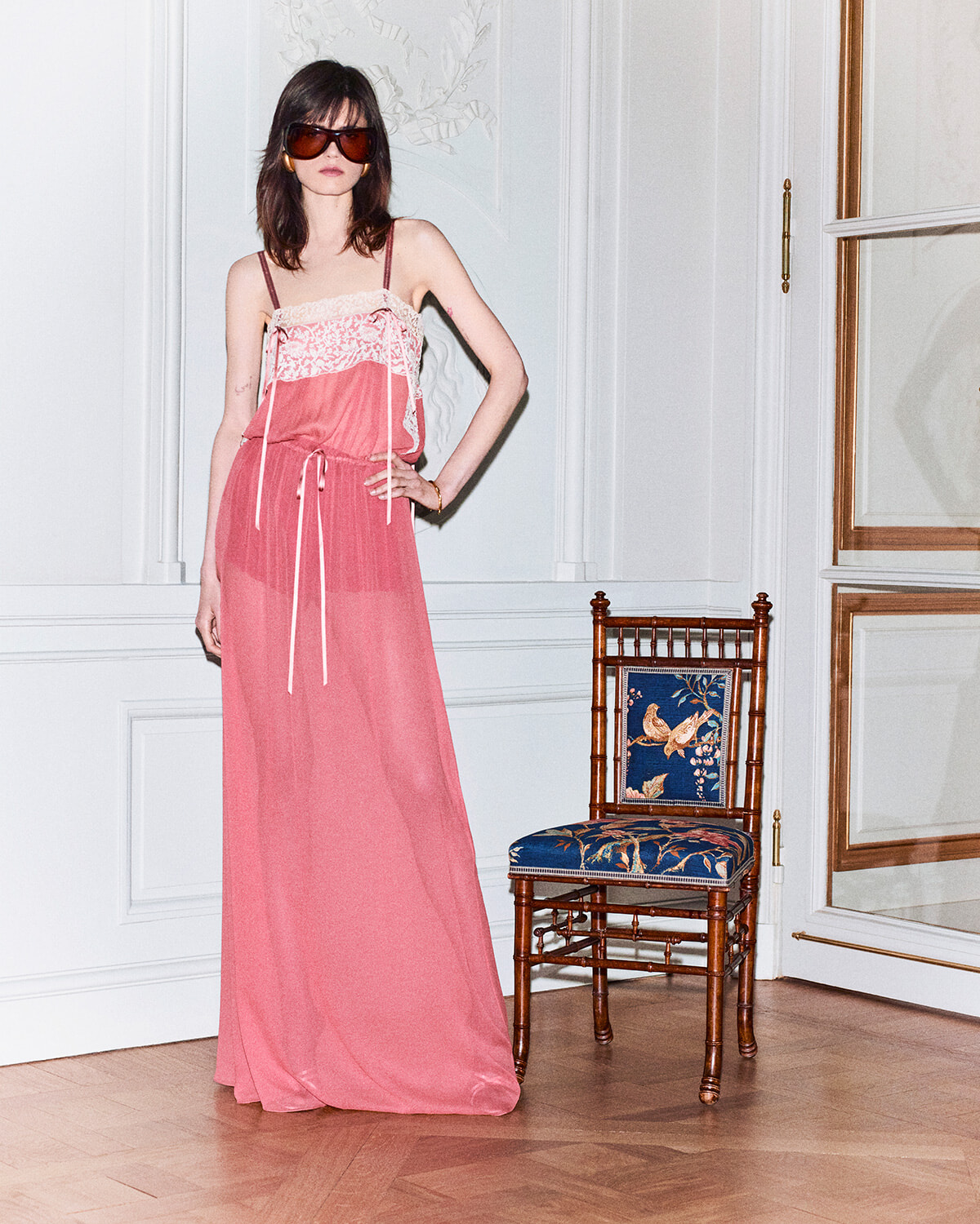 Model in pink lace-trim spaghetti-strap maxi dress with oversized sunglasses posing in a white-paneled room with a wooden chair nearby.