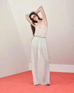 Woman in a sheer white maxidress standing on a pink carpet, arms raised, wearing sunglasses.