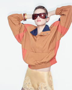 Model wearing an oversized orange jacket with blue collar, posing with hands behind head and futuristic sunglasses.