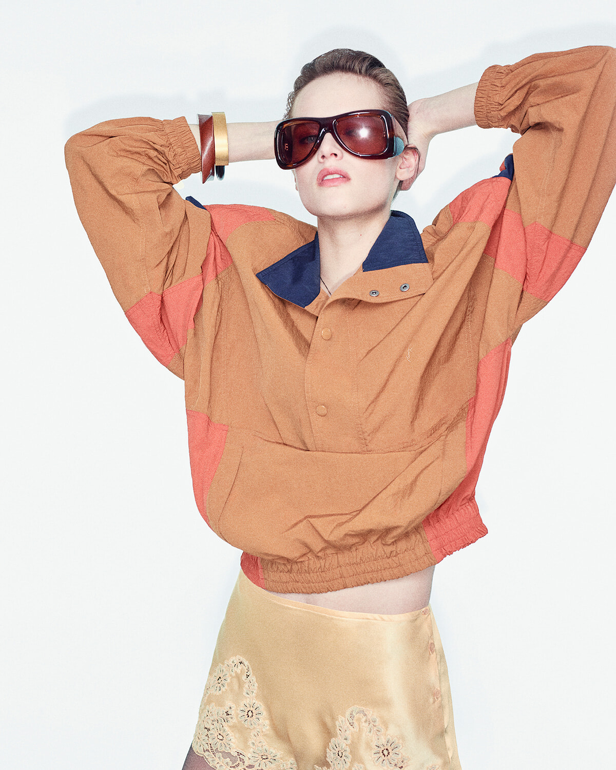 Model wearing an oversized orange jacket with blue collar, posing with hands behind head and futuristic sunglasses.
