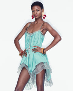 Model posing in a turquoise slip dress with lace trim, hands on hips, wearing large cherry earrings in a studio.