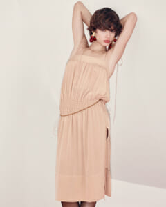 Model wearing a peach-midi dress with thin straps and a gold chain belt, arms raised behind her head, cherry earrings visible.