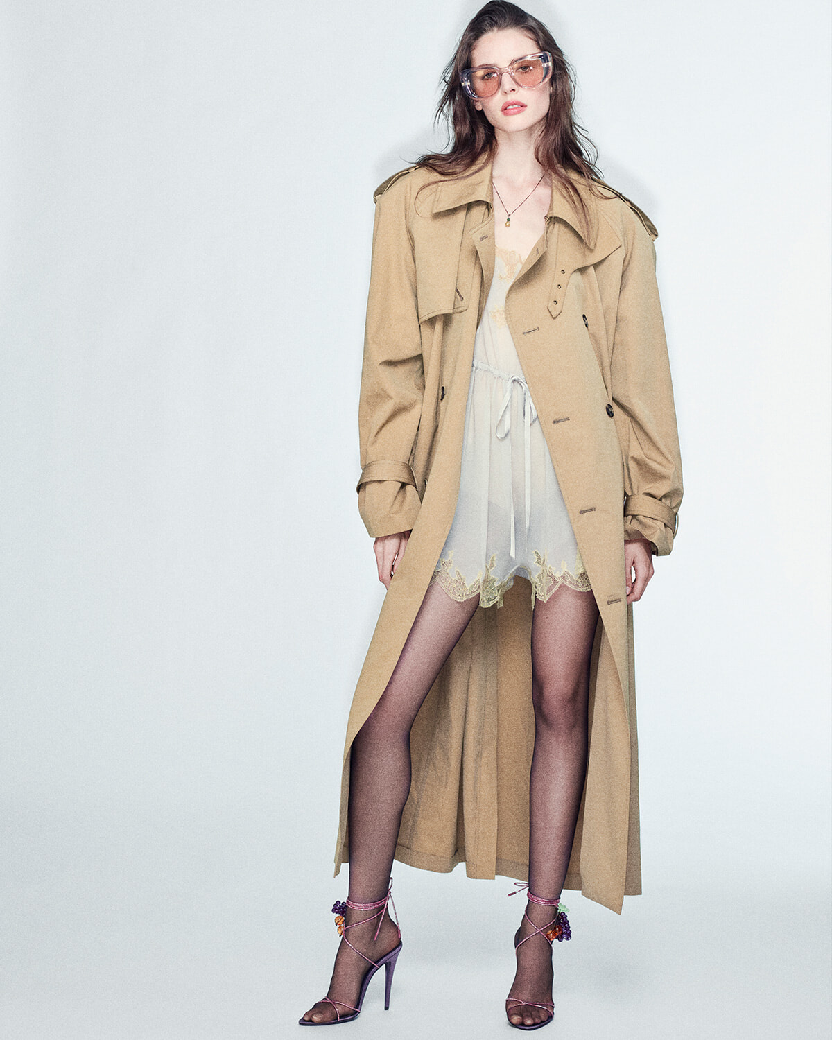 Female model wearing a tan trench coat over a sheer cream lace dress, pink sunglasses, and purple strappy heels, posing against a light gray background.