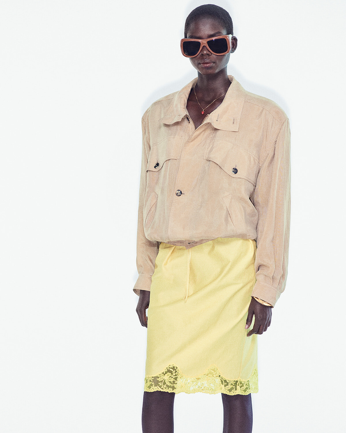 Model wearing a beige button-up jacket with a pale yellow lace-hem skirt and oversized brown sunglasses.
