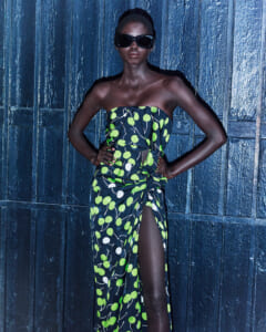 Dark-skinned model in a strapless lime-green leaf print dress with a high slit, hands on hips, wearing oversized black sunglasses against a blue wooden wall backdrop