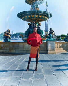 Person in a red jacket and color-block skirt poses in front of an ornate multi-tier fountain with statues and water jets in a sunny square.