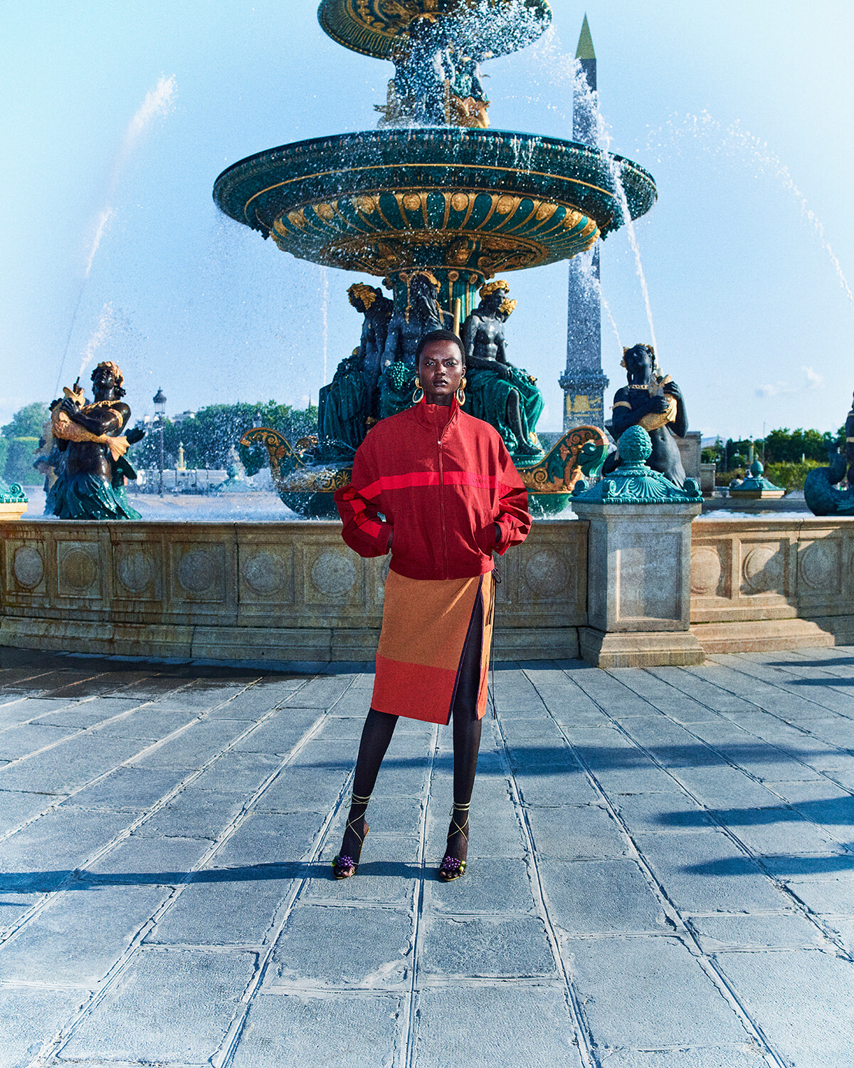 Person in a red jacket and color-block skirt poses in front of an ornate multi-tier fountain with statues and water jets in a sunny square.