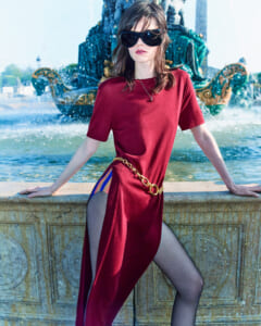 Woman in a burgundy dress with a high slit and gold chain belt, posing by an ornate fountain wearing oversized sunglasses.