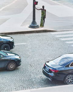 Woman in a strapless yellow-and-green patterned dress stands by a lamppost on a cobblestone urban street with parked cars nearby and a crosswalk in view.