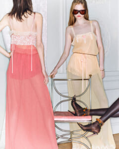 Two women model sheer pastel gowns in a vintage room; one faces away in a pink dress, the other wears a yellow dress with sunglasses standing by a chair.