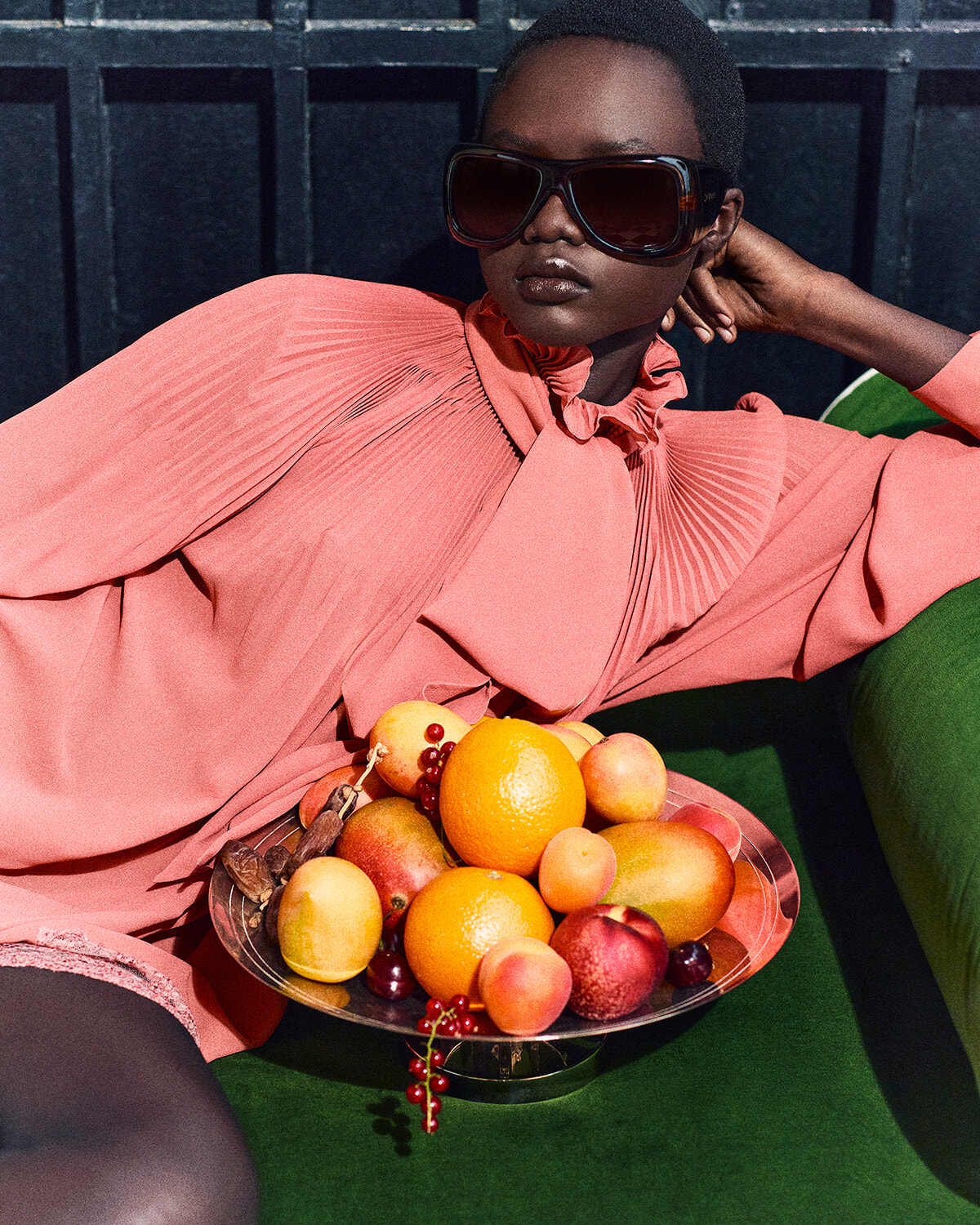 Model in coral pink pleated dress and oversized sunglasses reclines on a green sofa, with a silver fruit bowl of oranges, peaches, and berries nearby.