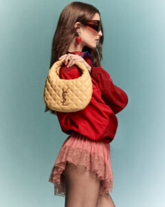 Model in a red jacket carries a beige quilted YSL shoulder bag, with pink lace skirt and oversized sunglasses in profile.