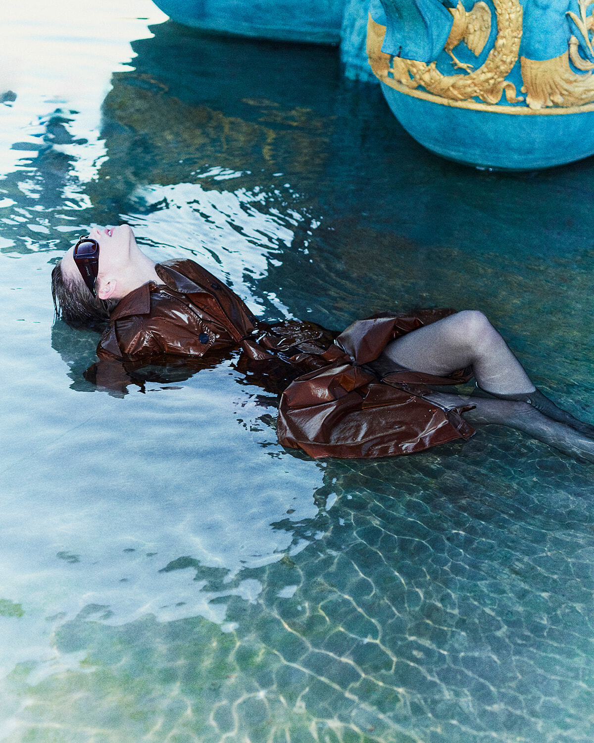 Person wearing a brown shiny coat and sunglasses floats on her back in clear water beside a blue and gold sculpture.