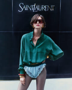 Model in a green oversized blouse and lace-trimmed underwear poses in front of a Saint Laurent sign, wearing large sunglasses and gold jewelry.