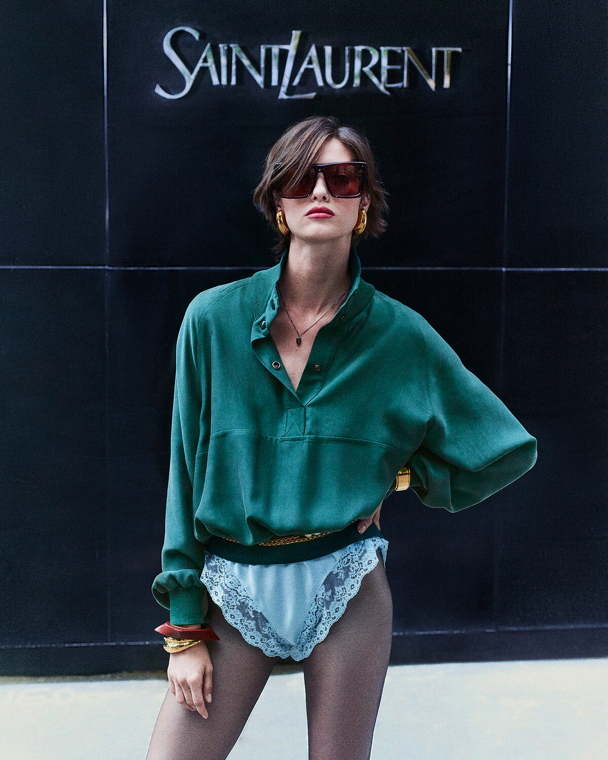 Model in a green oversized blouse and lace-trimmed underwear poses in front of a Saint Laurent sign, wearing large sunglasses and gold jewelry.