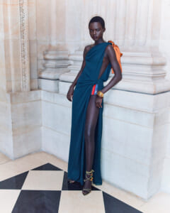 Model wearing a teal one-shoulder gown with a high slit, orange bag, and gold bracelets, standing against a stone column backdrop.