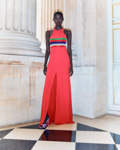 Model in a coral red sleeveless gown with rainbow waist stripes, standing beside marble columns on a black-and-white checkered floor.
