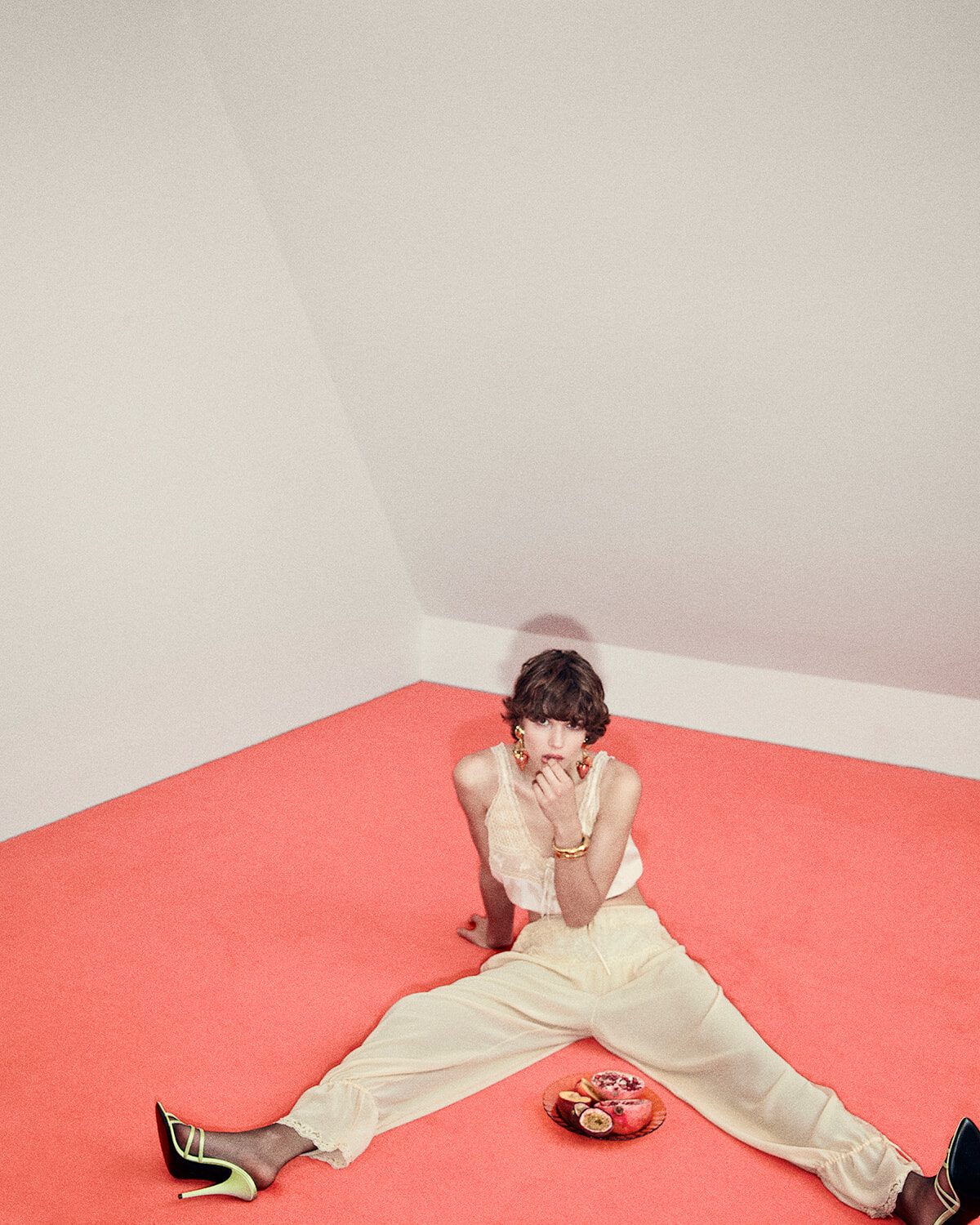 Model in a cream outfit sits on a pink floor with legs spread, neon-green heels, and a plate of fruit nearby in a minimal room.