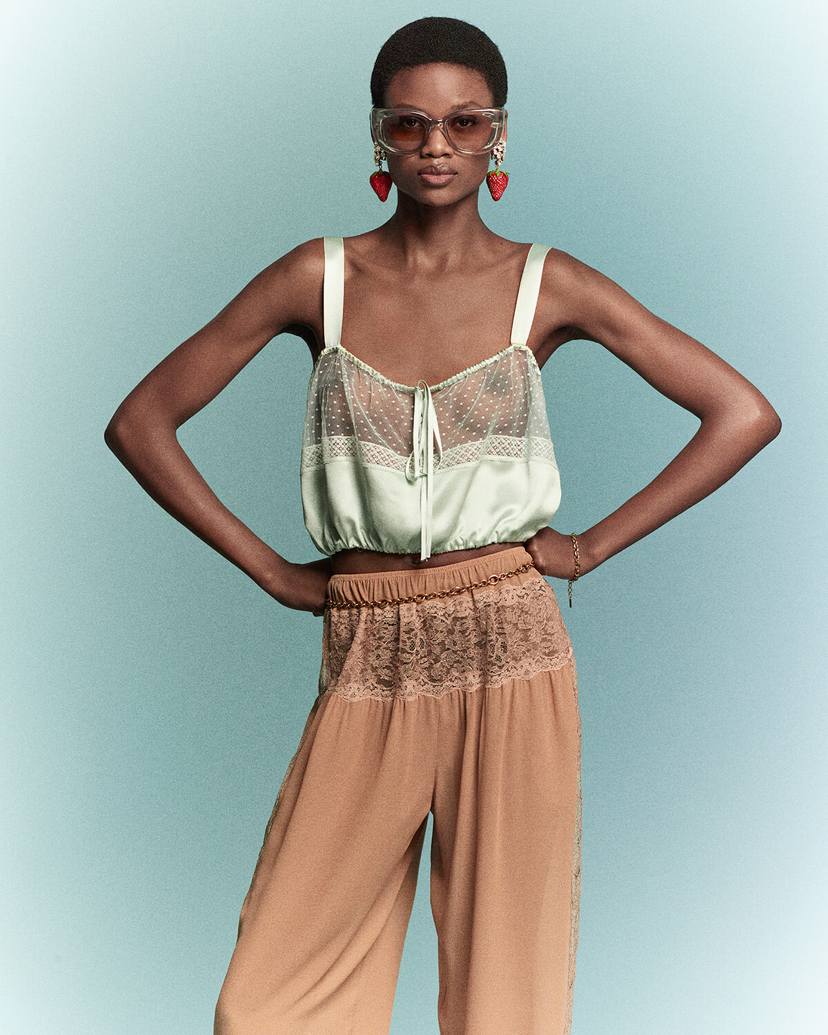 Model wearing a mint-green sheer lace top with satin straps and beige lace-trim wide-leg pants, posing with hands on hips, against a pale blue background with oversized sunglasses and strawberry earrings.