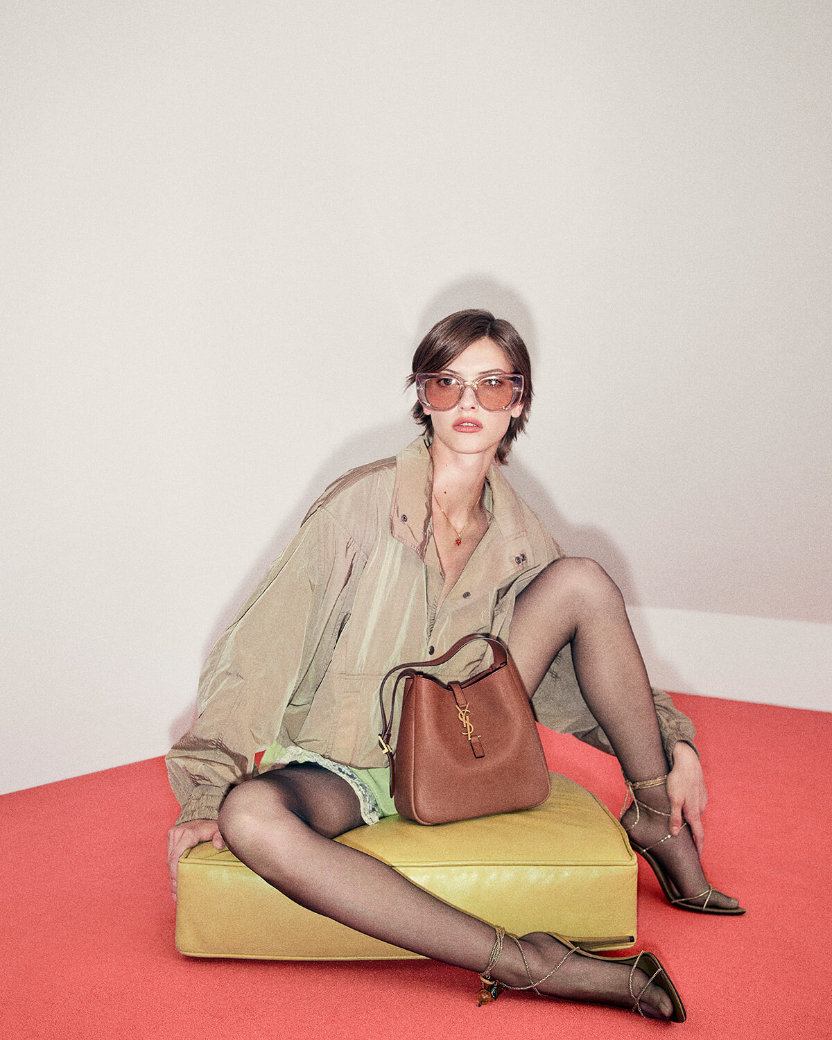 Model seated on a yellow ottoman wearing a beige windbreaker, sheer tights, and strappy sandals with a brown YSL handbag on her lap, stylish sunglasses on.