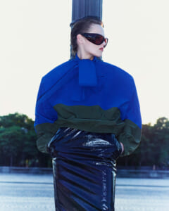 Model wearing an oversized cobalt blue and green color-blocked jacket with a shiny dark skirt, red-tinted sunglasses, leaning against a blue pillar outdoors.