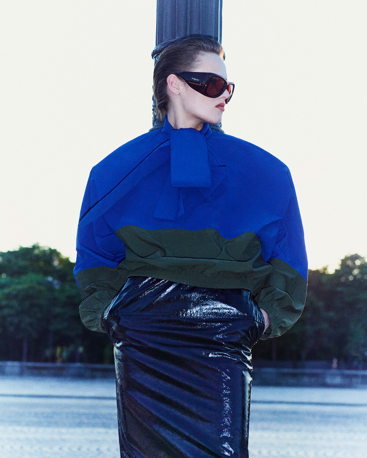 Model wearing an oversized cobalt blue and green color-blocked jacket with a shiny dark skirt, red-tinted sunglasses, leaning against a blue pillar outdoors.