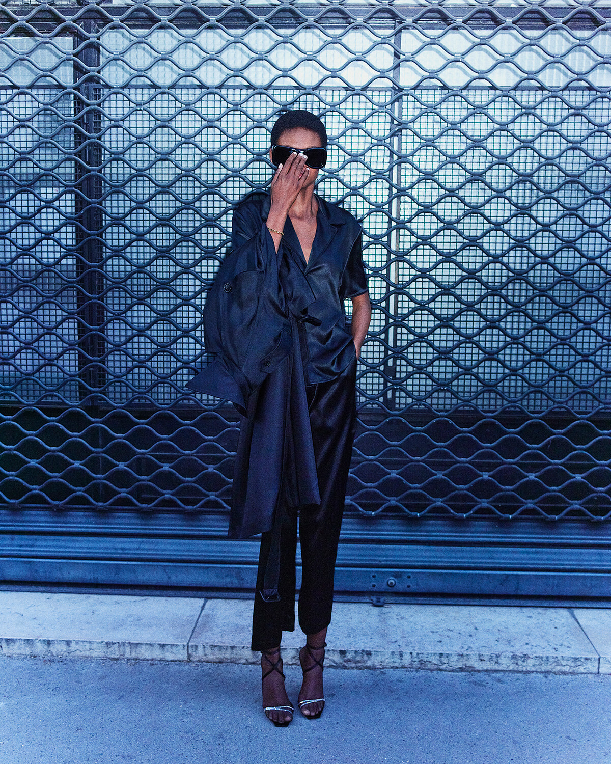 Person in a black satin outfit and oversized sunglasses poses against a metal lattice backdrop with one hand covering part of their face.