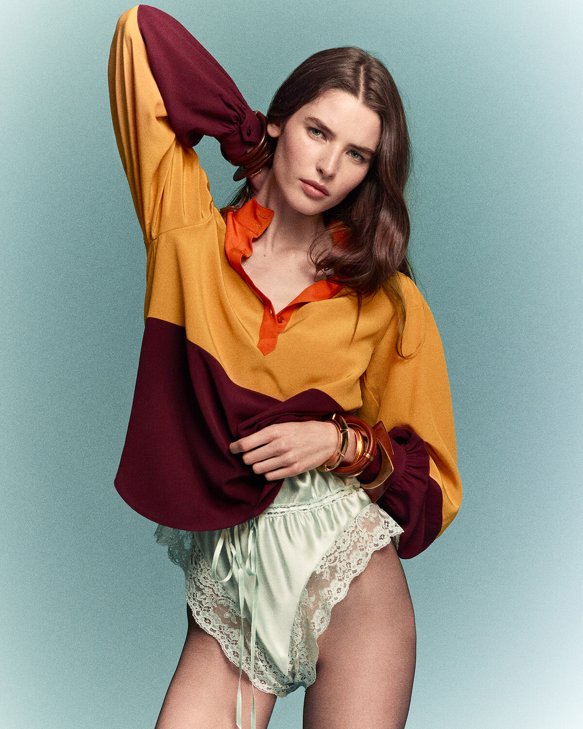 Model in a loose color-block blouse (mustard, orange, burgundy) with mint lace-trim shorts, posing with arm up against a teal backdrop.