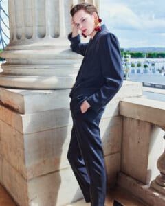 Person in a navy suit leaning against a large stone column on a balcony, wearing bold red cherry earrings, with an urban plaza in the background.