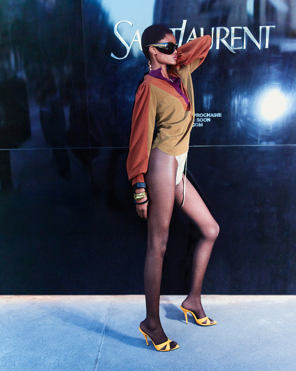 Model posing in a tan and orange color-block blouse, sheer tights, and yellow high-heeled sandals against a Saint Laurent backdrop.