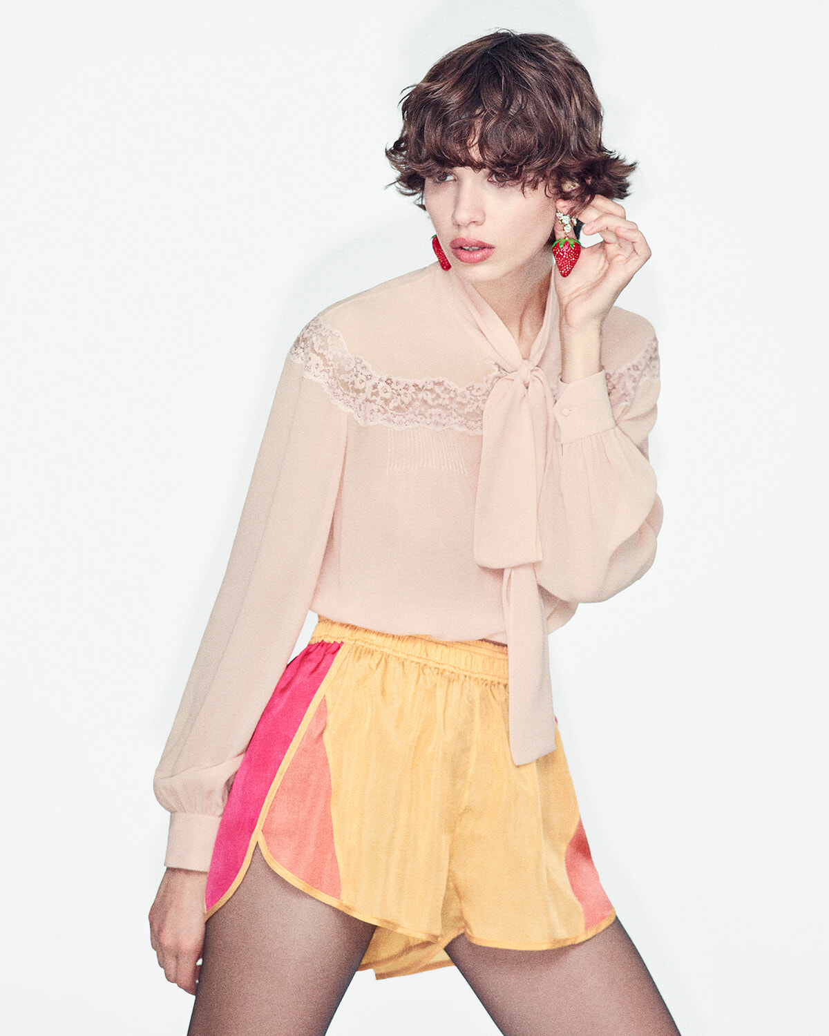 Model wearing a pale pink lace-yoked blouse with a neck bow, and color-blocked pink and yellow shorts; holding a strawberry-shaped earring nearby.