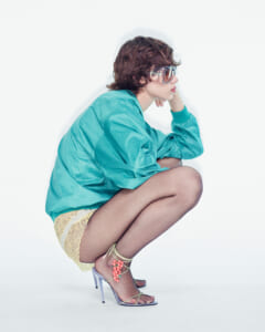 Person crouching in a turquoise jacket, lacy pale shorts, and strappy heels with pink bead accents, wearing oversized clear goggles/glasses.