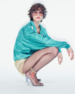 Model crouching in a turquoise jacket, lace shorts, and strappy high heels against a white backdrop with oversized sunglasses.