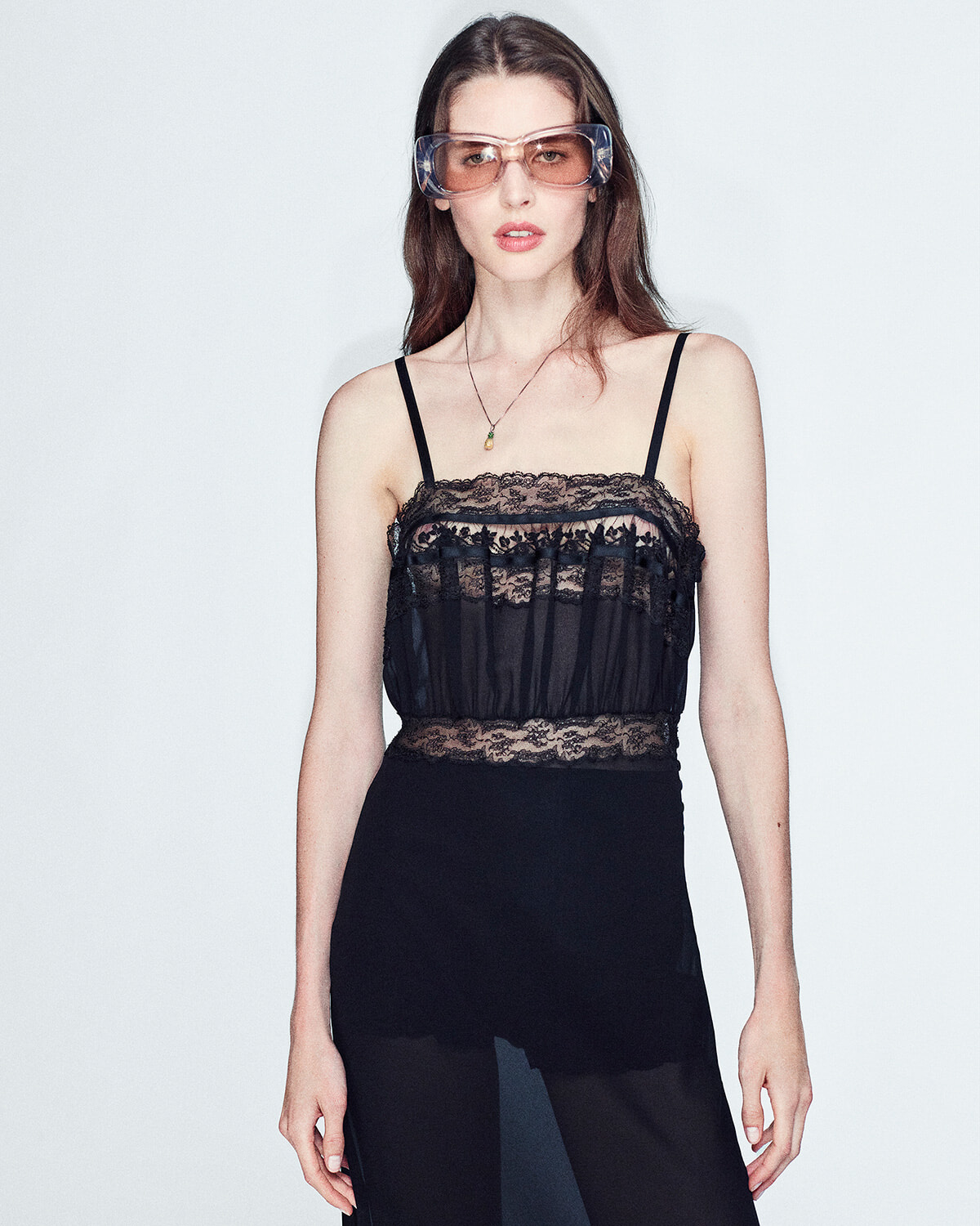 Model wearing a black lace camisole and sheer black pants with thin straps and oversized pink-tinted safety glasses lingeringly posing against a light background.