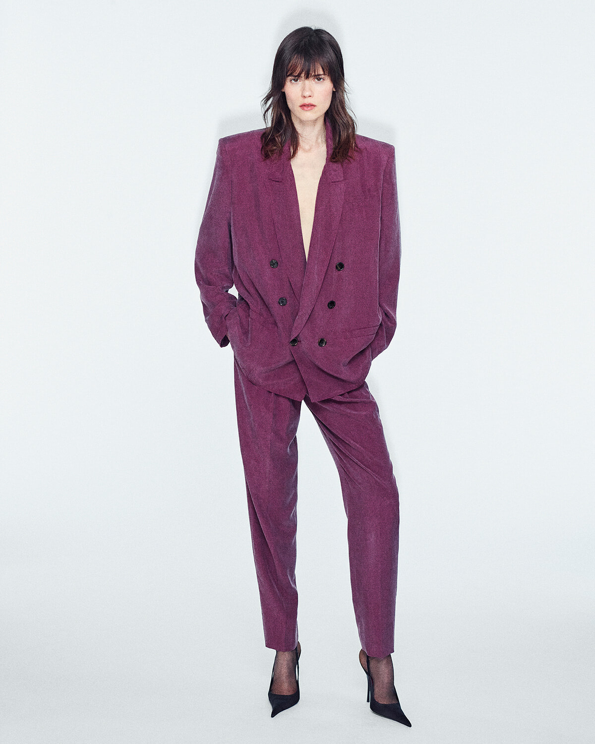 Woman in a magenta velvet double-breasted suit with hands in pockets, standing against a light background.