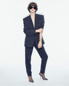 Person in a dark oversized suit with blazer and pants, wearing large black sunglasses and gold bracelets in a studio shot, posing confidently with one leg forward.