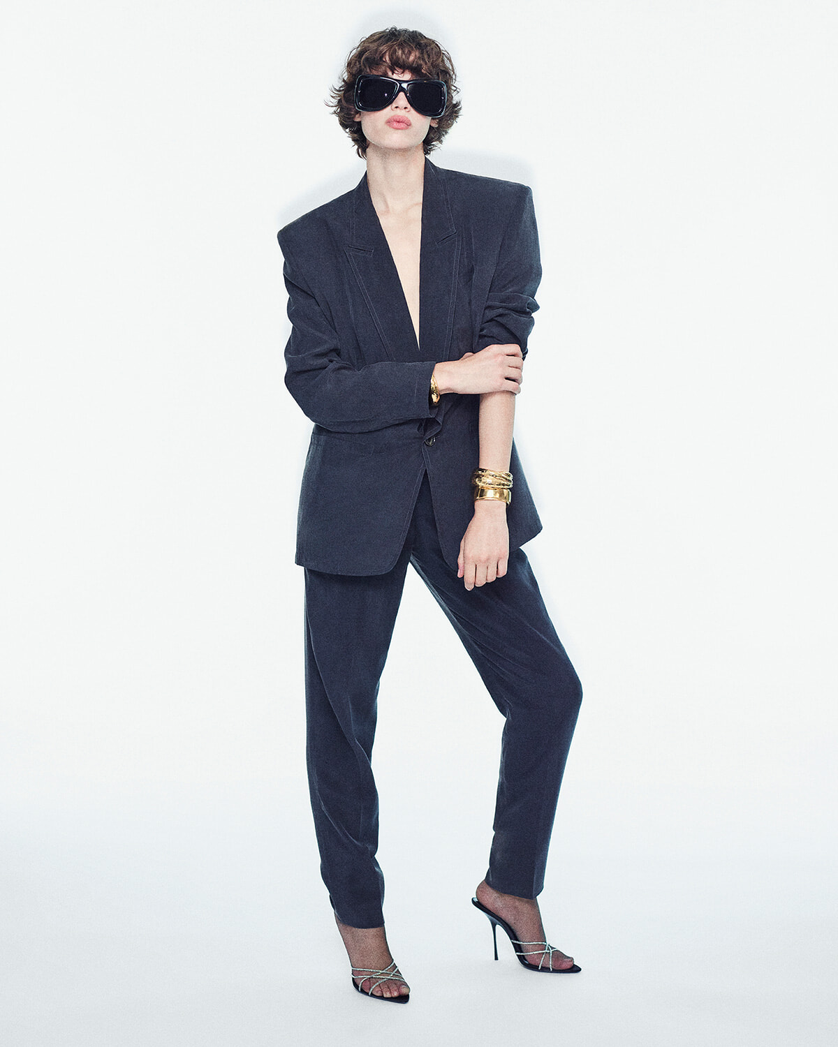 Person in a dark oversized suit with blazer and pants, wearing large black sunglasses and gold bracelets in a studio shot, posing confidently with one leg forward.