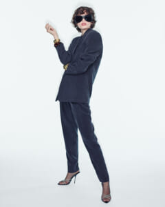 Fashion model in a navy velvet suit with oversized sunglasses, gold bangles, and strappy heels posing against a white background.