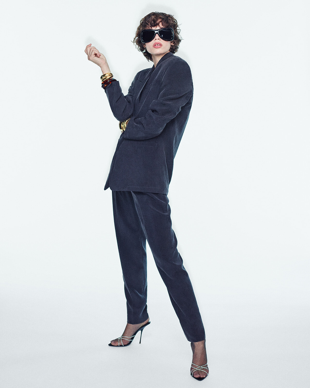 Fashion model in a navy velvet suit with oversized sunglasses, gold bangles, and strappy heels posing against a white background.