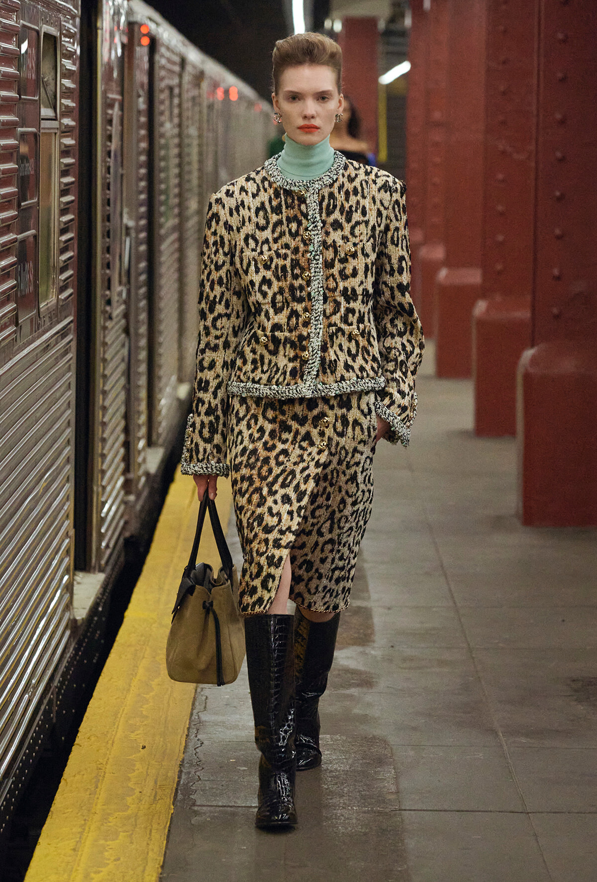 Woman in a leopard‑print matching suit walks on a subway platform, holding a beige bag, wearing a turquoise turtleneck and black knee‑high boots.