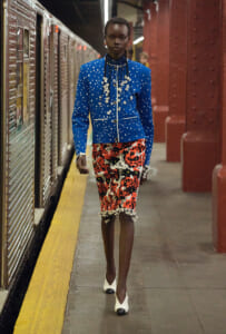 Model walking on a subway platform in a cobalt blue, polka-dotted jacket and red floral skirt with white heels and floral jewelry detail.
