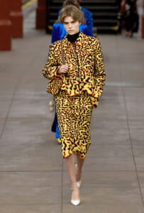 Model walking a runway in a yellow-orange animal-print suit with black accents, carrying a patterned bag and wearing a black turtleneck and white heels.
