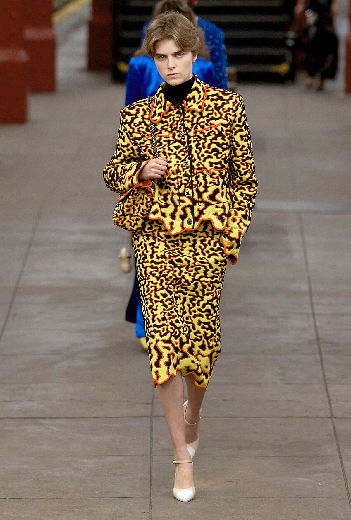 Model walking a runway in a yellow-orange animal-print suit with black accents, carrying a patterned bag and wearing a black turtleneck and white heels.