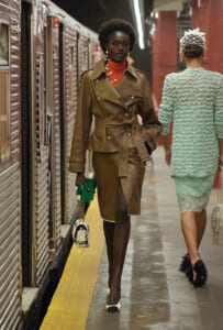 Two models walk down a subway platform; foreground model wears a brown belted trench coat, red turtleneck, green gloves, and a patterned handbag.