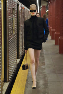 Woman in a black tailored jacket and miniskirt with fringe walks along a subway platform, oversized sunglasses in place.