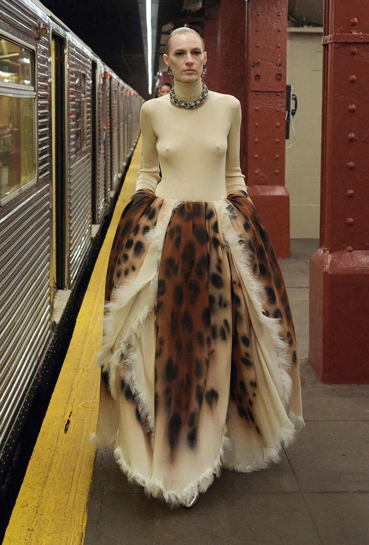 Woman in a beige turtleneck and voluminous leopard-print skirt walks on a subway platform, accessorized with a chunky necklace and earrings.