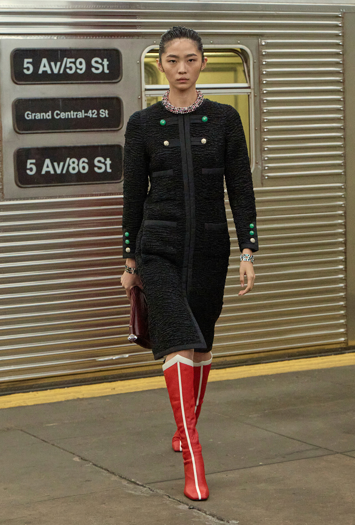 Model walking on a subway platform in a black textured coat with green and gold buttons, holding a burgundy clutch, and red knee-high boots with white trim.