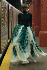 Model in a black fitted top and a voluminous teal-green patterned skirt with white feather trim, accessorized with a bold green necklace and sunglasses in a subway station.