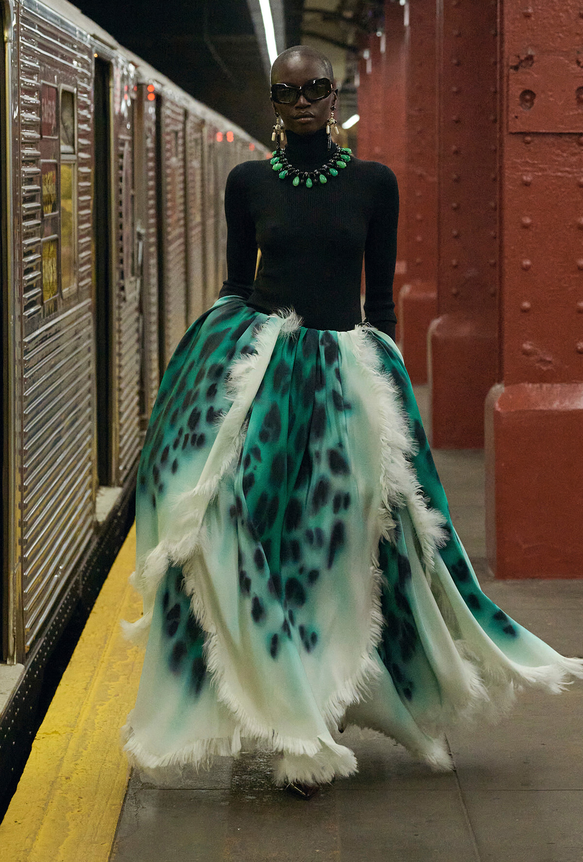 Model in a black fitted top and a voluminous teal-green patterned skirt with white feather trim, accessorized with a bold green necklace and sunglasses in a subway station.