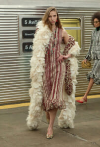 Model wearing a red and ivory striped midi dress with a long cream feathered coat on a subway platform, gold heels visible.